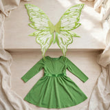 Translucent fairy wings with lime green patterns lie above a green long-sleeved dress on a cream surface beside a window.