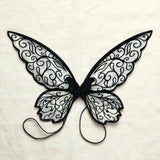 Translucent fairy wings with black swirling patterns and two black shoulder loops lie flat on cream fabric.
