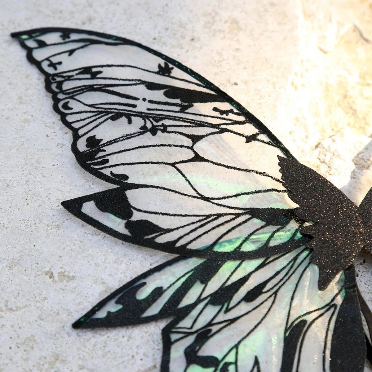 A close-up of iridescent fairy wings shows translucent fabric with black glitter outlines and vein patterns on a pale textured surface.