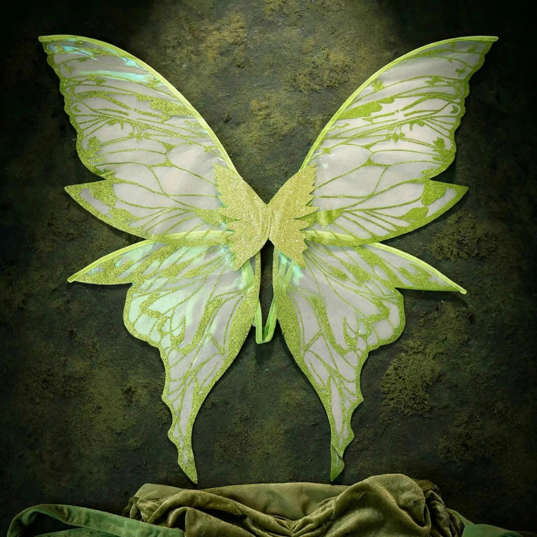 Pale iridescent fairy wings with lime green glitter patterns hang against a dark textured wall above draped green fabric.