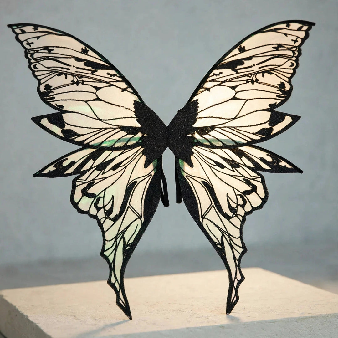 Iridescent fairy wings with black glitter vein patterns and long pointed lower tips stand on a cream pedestal against a gray background.