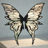 Iridescent fairy wings with black glitter vein patterns and long pointed lower tips stand on a cream pedestal against a gray background.