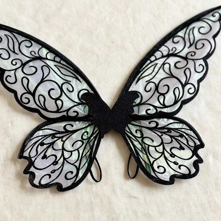Iridescent fairy wings with black swirling patterns and shoulder loops rest on cream fabric.