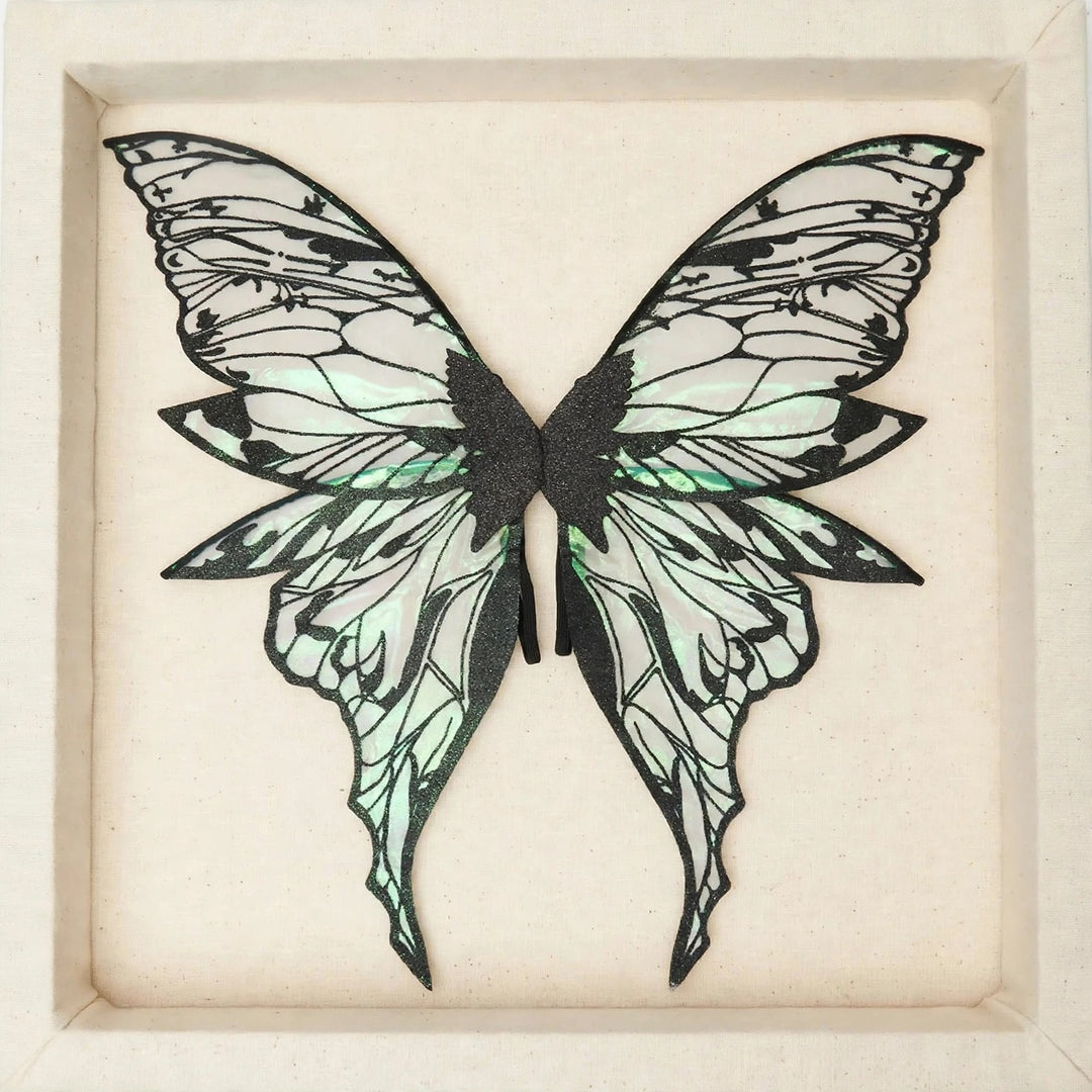 Translucent fairy wings with pale green reflections, black glitter patterns, and pointed lower tips lie within a recessed cream panel.