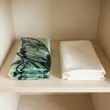 Folded pale green fairy wings with black outlines sit beside folded white fabric on a cream cupboard shelf.