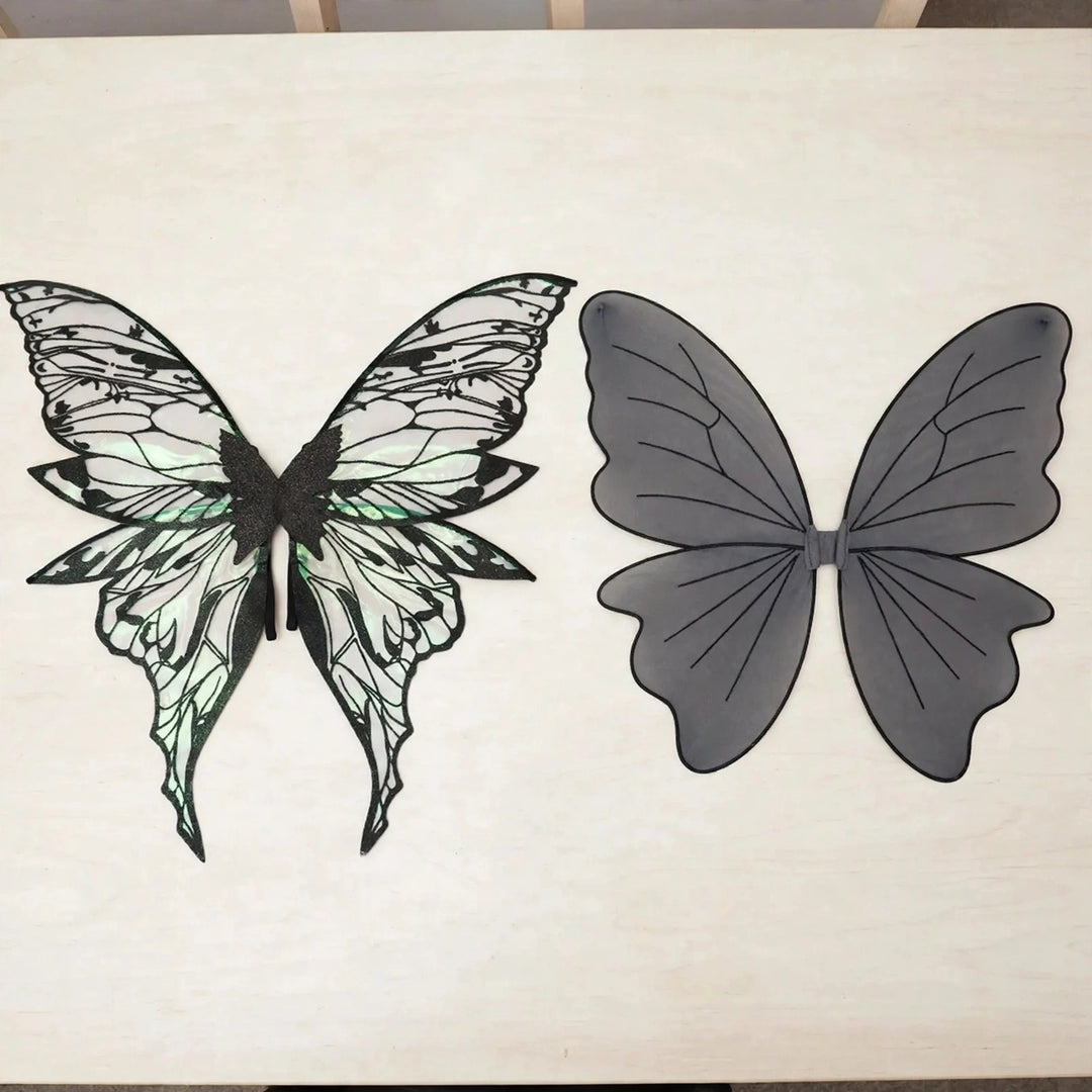 Iridescent fairy wings with intricate black patterns lie beside gray fairy wings with simple black veins on a white tabletop.