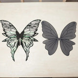 Iridescent fairy wings with intricate black patterns lie beside gray fairy wings with simple black veins on a white tabletop.