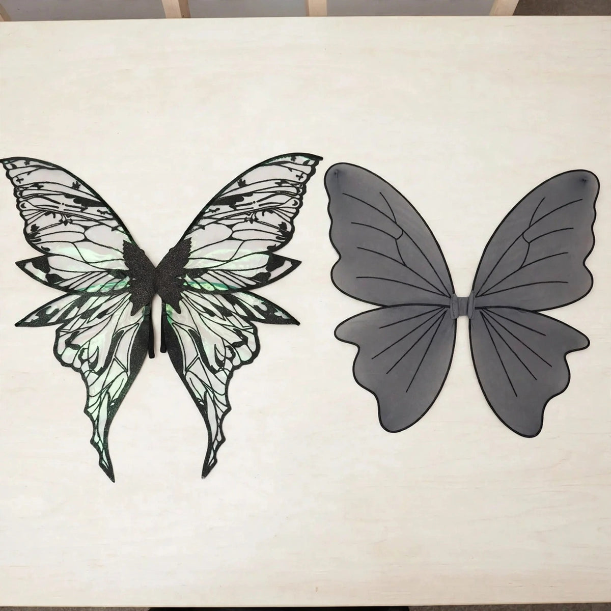 Iridescent fairy wings with intricate black patterns lie beside gray fairy wings with simple black veins on a white tabletop.