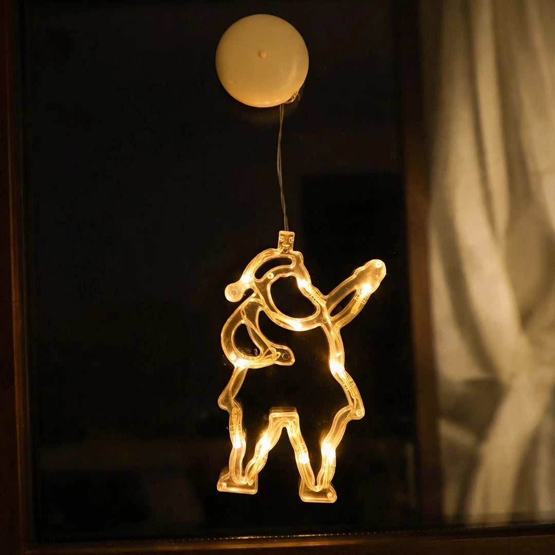 Clear Santa-shaped lighted ornament glows with warm white lights on a dark window beside a pale curtain, beneath a round cream suction cup.