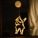Clear Santa-shaped lighted ornament glows with warm white lights on a dark window beside a pale curtain, beneath a round cream suction cup.