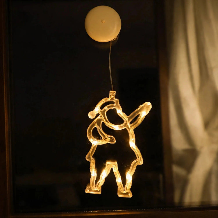 Clear Santa-shaped lighted ornament glows with warm white lights on a dark window beside a pale curtain, beneath a round cream suction cup.