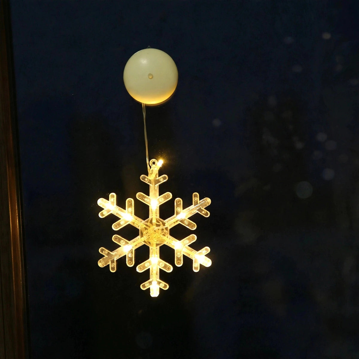 A clear snowflake-shaped lighted ornament glows warm yellow beneath a round cream housing against a dark window.