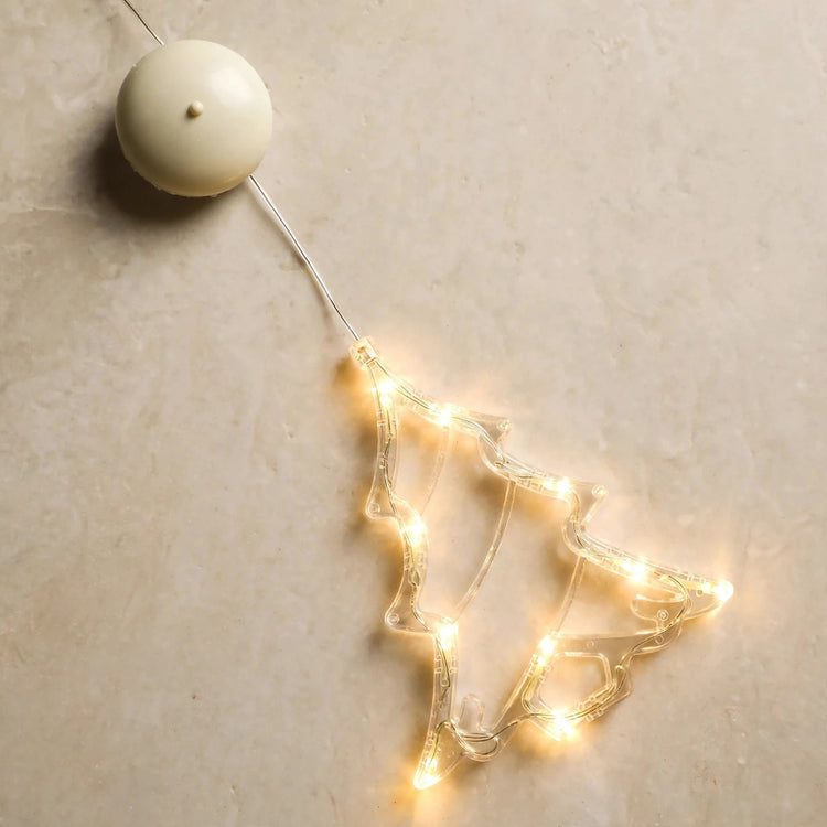 Clear tree-shaped lighted ornament with warm white lights lies diagonally on a beige surface, wired to a round cream battery housing.