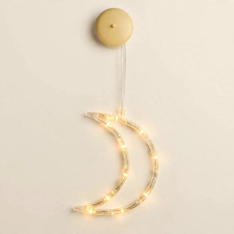 A clear crescent moon-shaped lighted ornament glows warm yellow beneath a round beige housing against an off-white background.