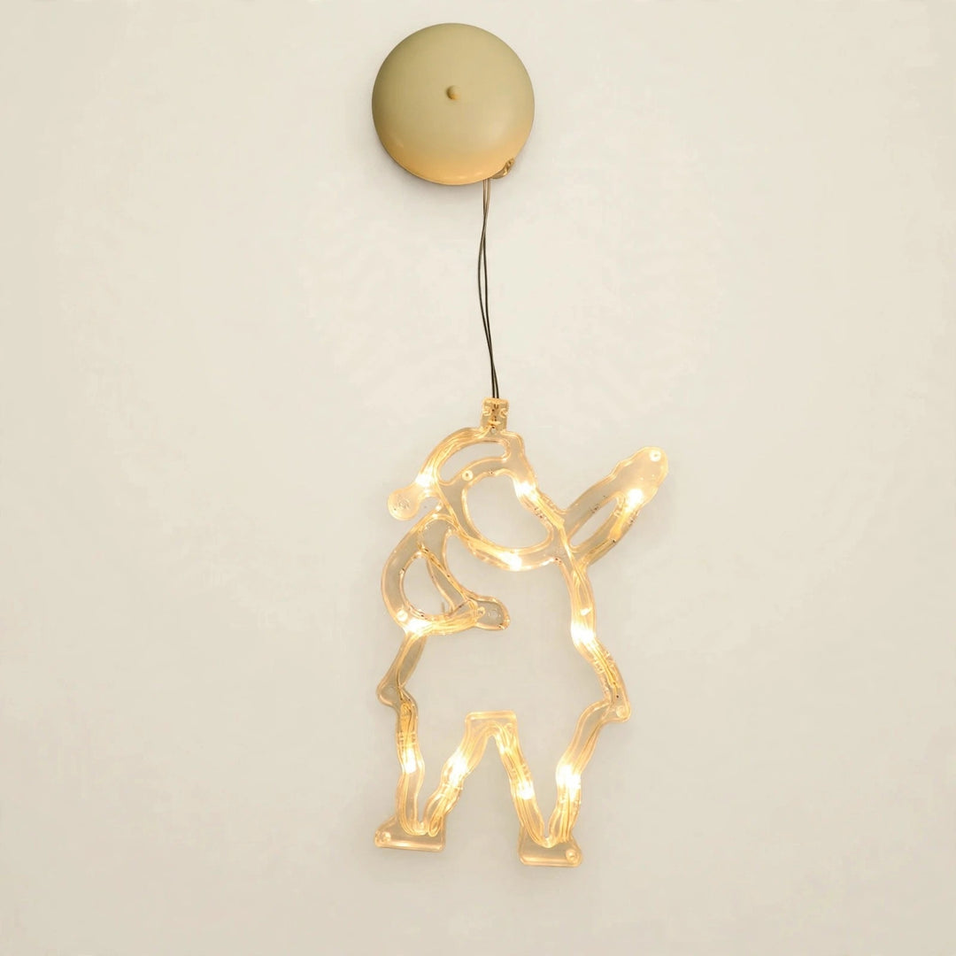 A clear Santa-shaped lighted ornament with one raised arm glows warm yellow beneath a round beige housing against an off-white background.