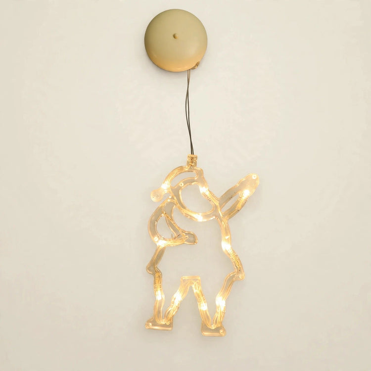 A clear Santa-shaped lighted ornament with one raised arm glows warm yellow beneath a round beige housing against an off-white background.