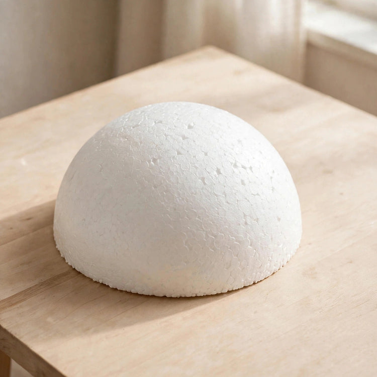 A white polystyrene foam half ball with visible bead texture rests on a pale wooden table beside a softly lit window.