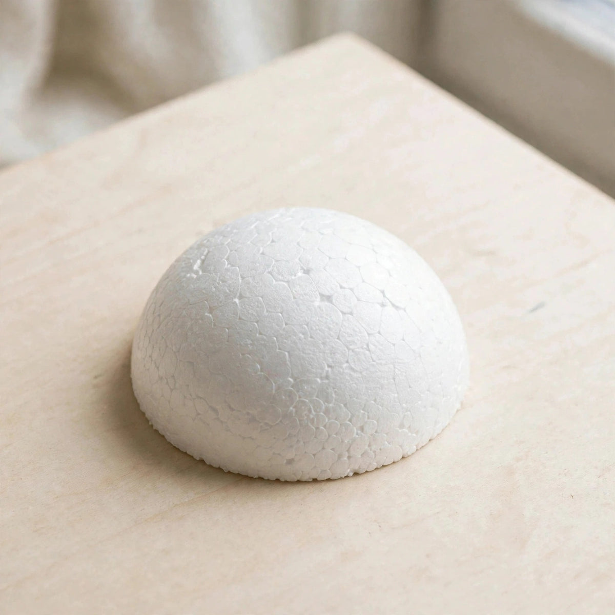 A white polystyrene foam half ball with visible bead texture rests on a light wooden tabletop, viewed diagonally from above.