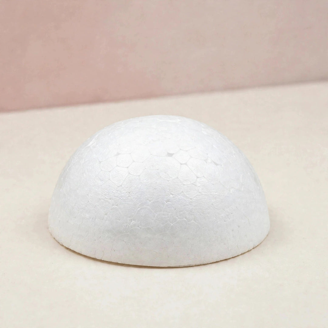 A white polystyrene foam half ball with a textured surface sits on a cream tabletop against a muted pink background.