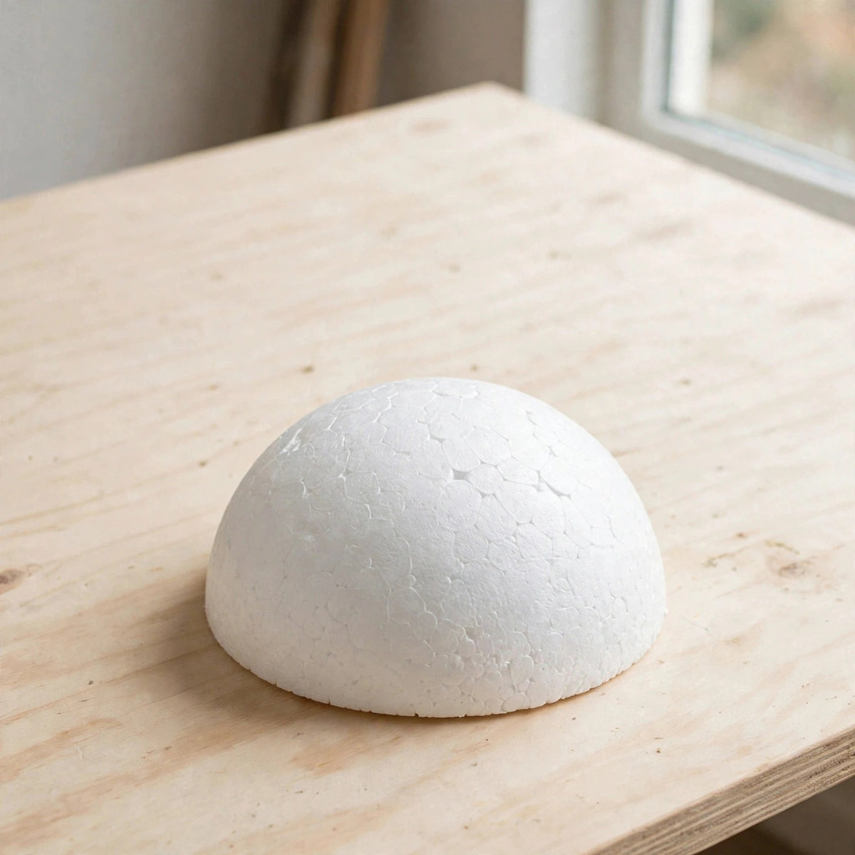 A white polystyrene foam half ball rests on a pale wooden table beside a window, with its textured surface visible.