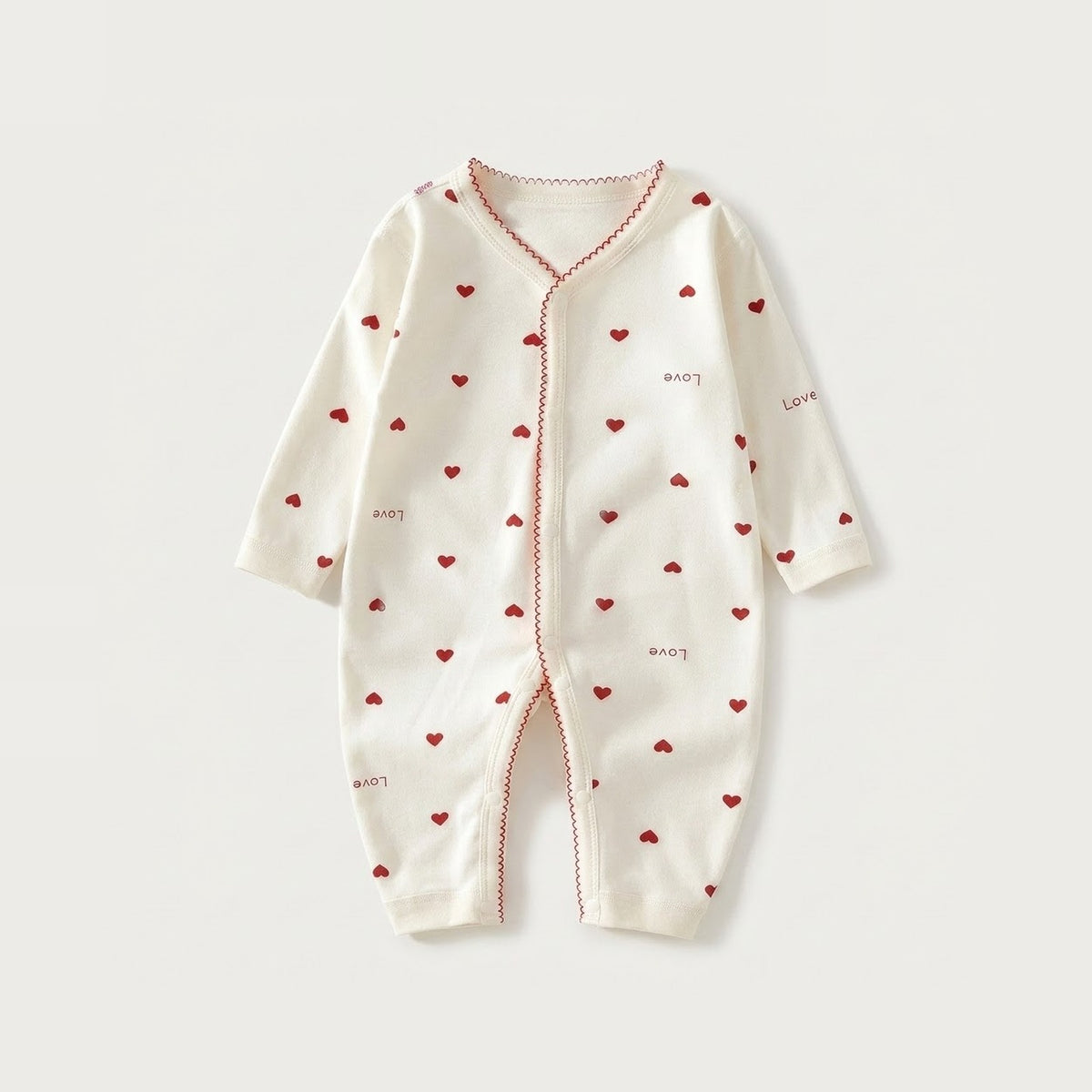 Cream long-sleeved baby romper with red hearts, scattered love lettering, and red scalloped trim lies flat on a pale background.