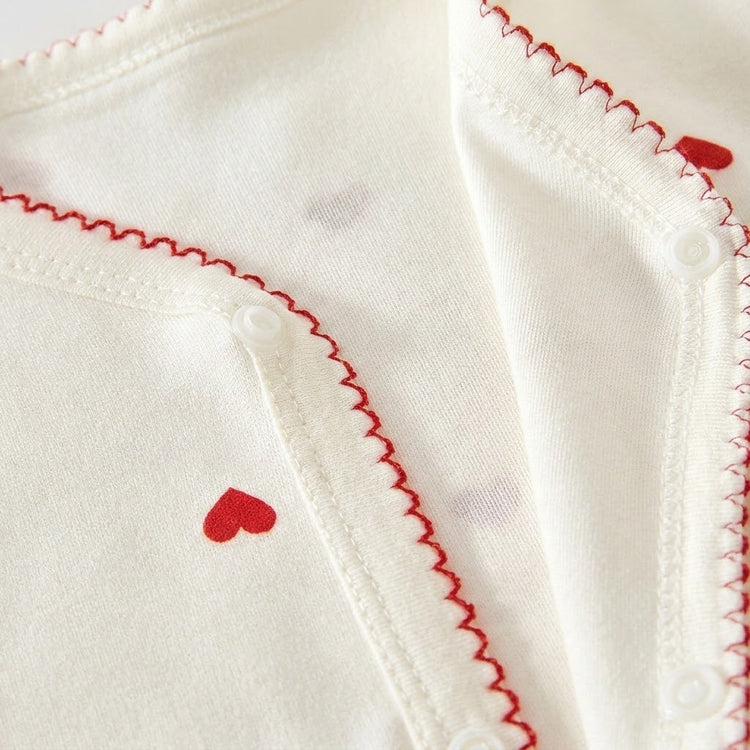 Close-up of a cream baby romper neckline showing red scalloped stitching, heart prints and white snap fasteners.