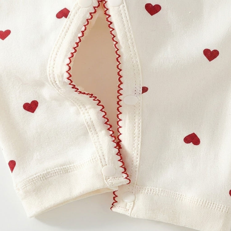 Cream baby romper legs show red heart prints, red zigzag trim, and an open snap-button inseam against a white background.