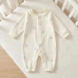 Cream ribbed baby romper with pale-blue motifs lies on a white knitted blanket beside white crib rails and light wooden flooring.