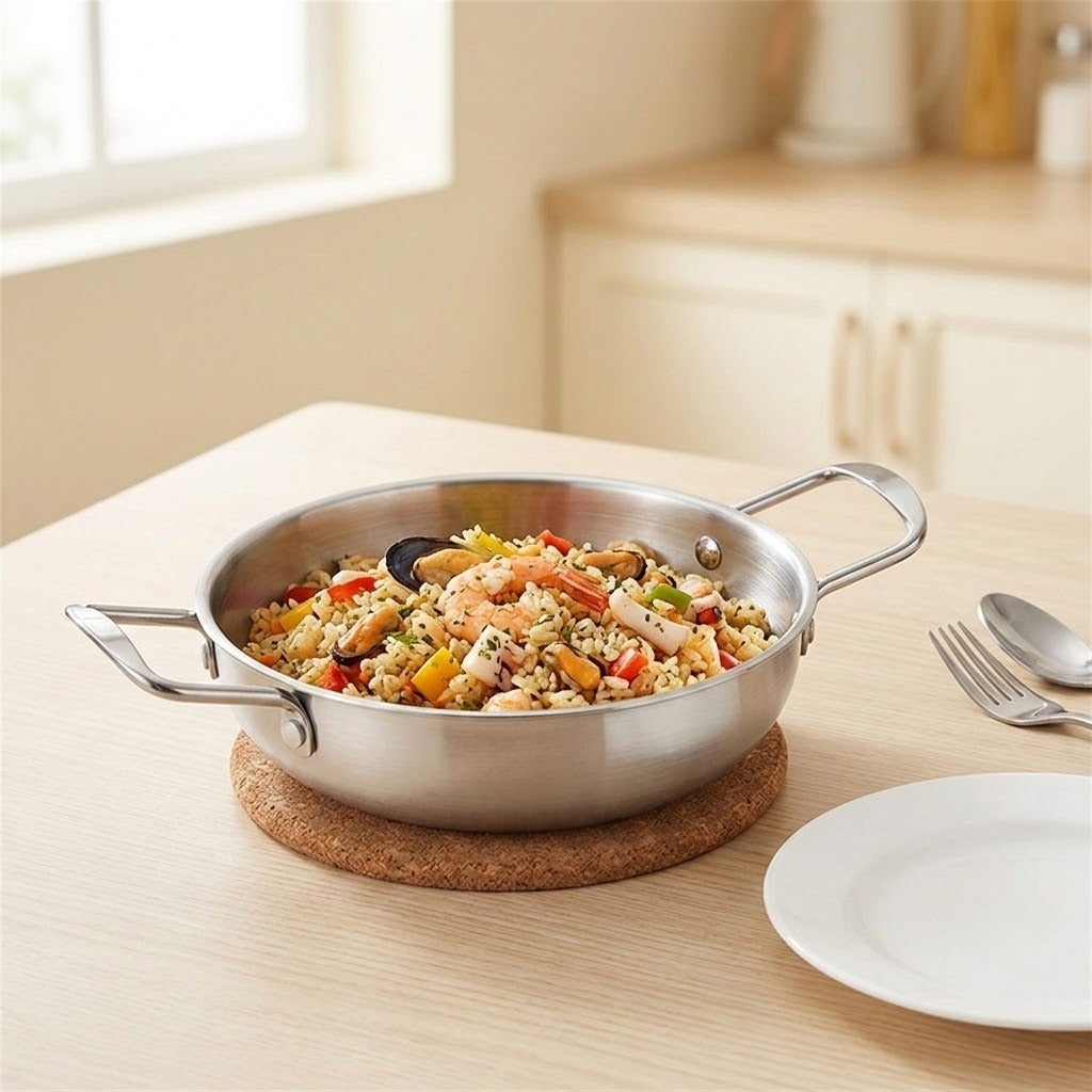 A stainless steel paella pan with two side handles, filled with a colorful seafood rice dish, sitting on a cork trivet on a light wood table in a bright kitchen setting.
