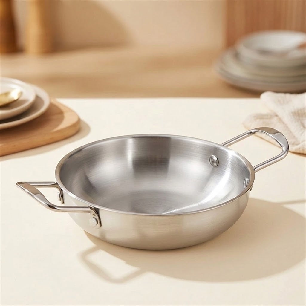 A stainless steel paella pan with two side handles sits on a light beige tabletop in a softly blurred kitchen.
