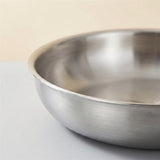 Close-up of a stainless steel paella pan showing its brushed exterior and rim against a warm beige background.