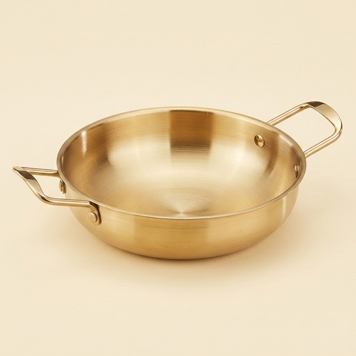 A gold-toned paella pan with riveted loop handles is shown close up on a cream background.