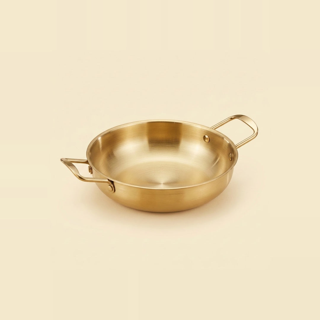 A gold-toned paella pan with two rectangular loop handles sits on a warm cream background.