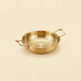 A gold-toned paella pan with two rectangular loop handles sits on a warm cream background.
