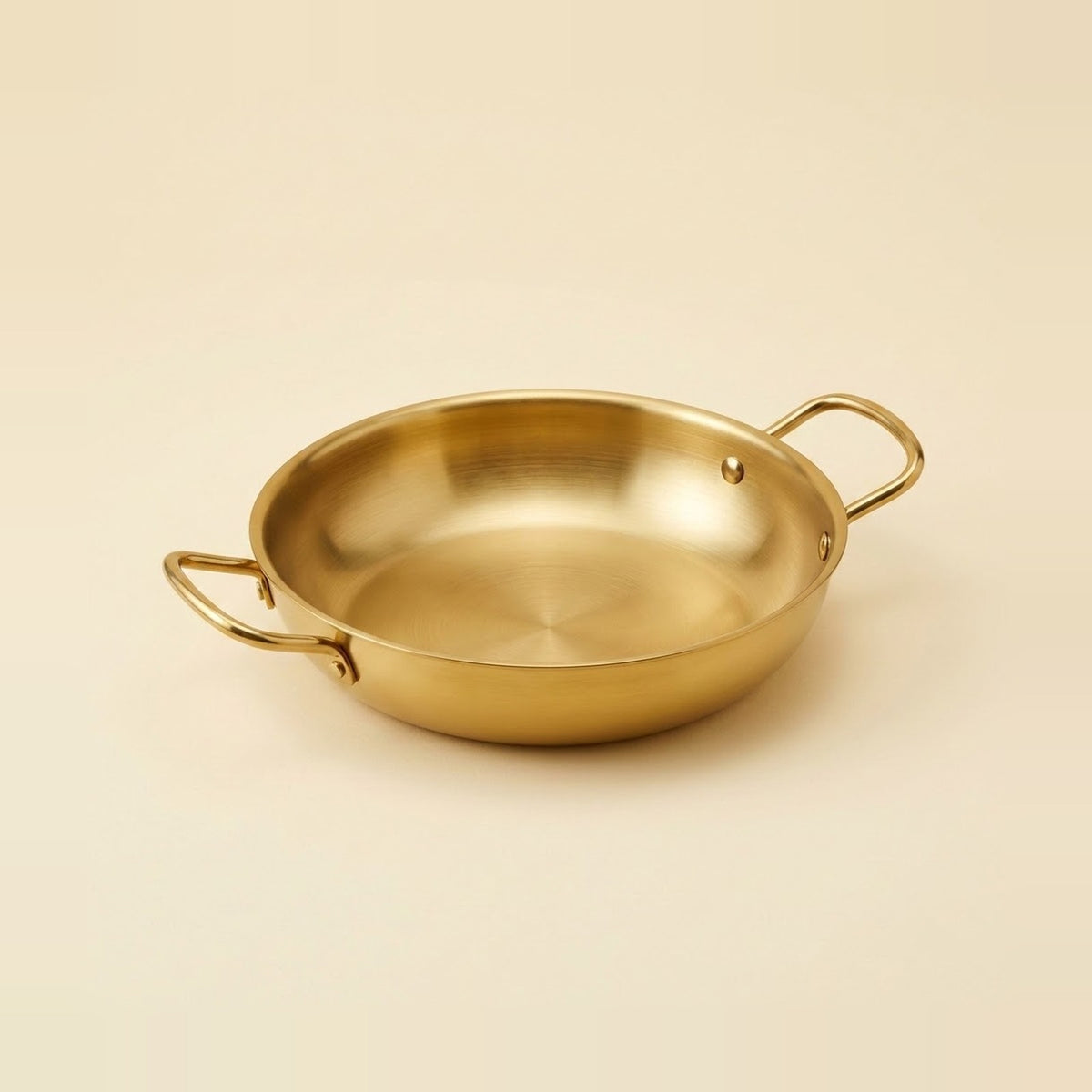 A gold-toned paella pan rests on a warm beige background, with its empty interior and two loop handles visible.