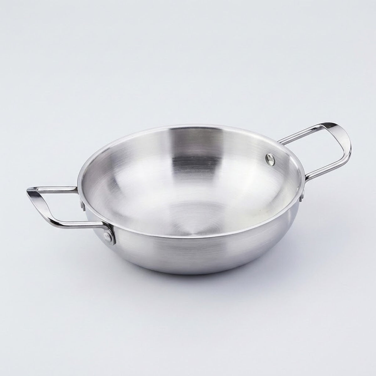 A silver stainless steel paella pan with riveted side handles is shown close up against a light gray background.