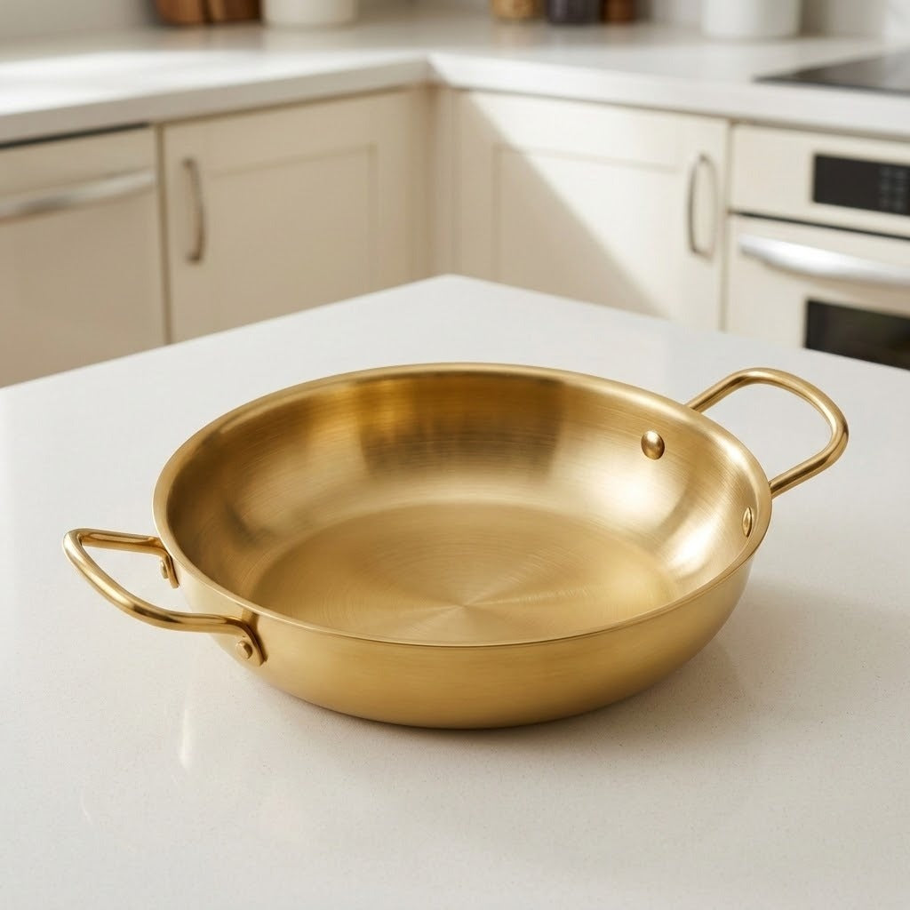 A gold-colored paella pan with two side handles sits empty on a white countertop in a softly blurred kitchen.