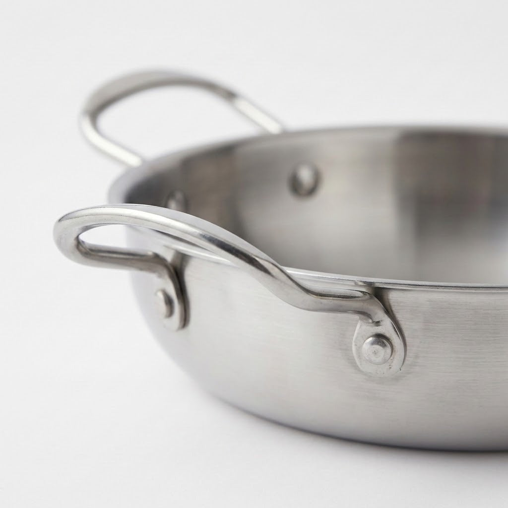 Close-up of a stainless steel paella pan highlighting its curved side handle and riveted attachments against a white background.