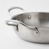 Close-up of a stainless steel paella pan highlighting its curved side handle and riveted attachments against a white background.