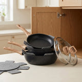 Black speckled cookware set with wood-look handles, glass covers, and gray pan protectors stacked on a counter beside an open wooden cabinet.