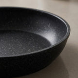Black cookware set detail showing a pan’s speckled non-stick interior and curved rim against a beige background.