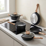 A set of black non-stick cookware with light wood handles and glass lids, arranged on a modern kitchen countertop next to a black induction cooktop. The set includes two pots, two pans, and a hanging pan against a tiled wall.
