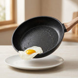 Black cookware set frying pan with a wood-look handle tilted to slide a fried egg onto a white plate.