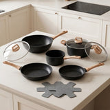 A 6-piece black non-stick cookware set with light wood handles and clear glass lids, arranged on a cream-colored kitchen countertop with matching cabinets in the background.