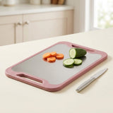 A rectangular cutting board with a dusty pink frame and a gray non-slip surface, shown on a kitchen counter with sliced carrots, cucumber pieces, and a small knife.