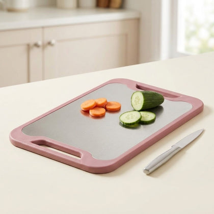 A rectangular cutting board with a dusty pink frame and a gray non-slip surface, shown on a kitchen counter with sliced carrots, cucumber pieces, and a small knife.