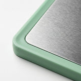 Close-up of a cutting board corner showing a brushed stainless steel surface and rounded mint green frame against white.