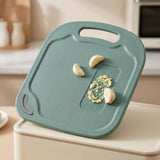 A teal-green plastic cutting board with a handle and a textured grater section, shown with garlic cloves on top in a softly blurred kitchen setting.