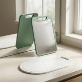 A set of three kitchen cutting boards on a light beige countertop near a window: two sage green boards standing upright, one lying flat, and a white oval board in the foreground.