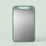 A rectangular cutting board with a brushed silver metal center and a sage green border, standing upright against a light mint green background. The brand name 'Bocle' is printed near the bottom.